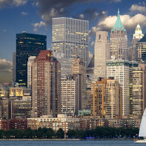 Panoramic view of New York City skyline with sailboat and river during twilight.