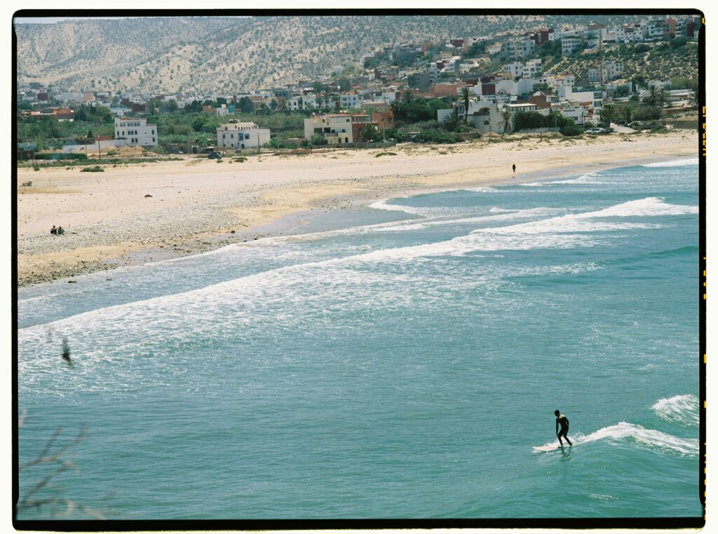 Aerial view of a surfer catching waves along the scenic Moroccan beach with mountains in the background.
