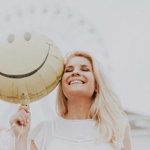 Cheerful woman holding a smiley balloon outdoors on a sunny day, exuding happiness and positivity.