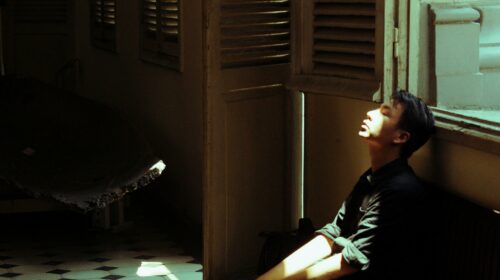A young man sits in a dimly lit room, soaked in sunlight, depicting introspection.