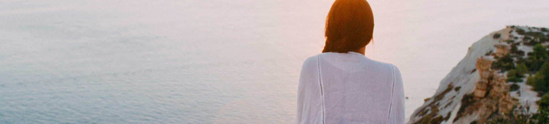 A serene view of a woman sitting on a rocky cliff, gazing at the ocean during sunset. Perfect for relaxation and travel themes.