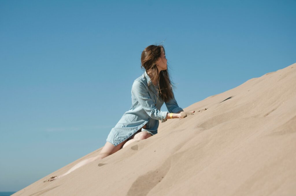 Woman in casual attire climbing sand dunes under a clear blue sky in Morocco.