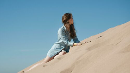 Woman in casual attire climbing sand dunes under a clear blue sky in Morocco.
