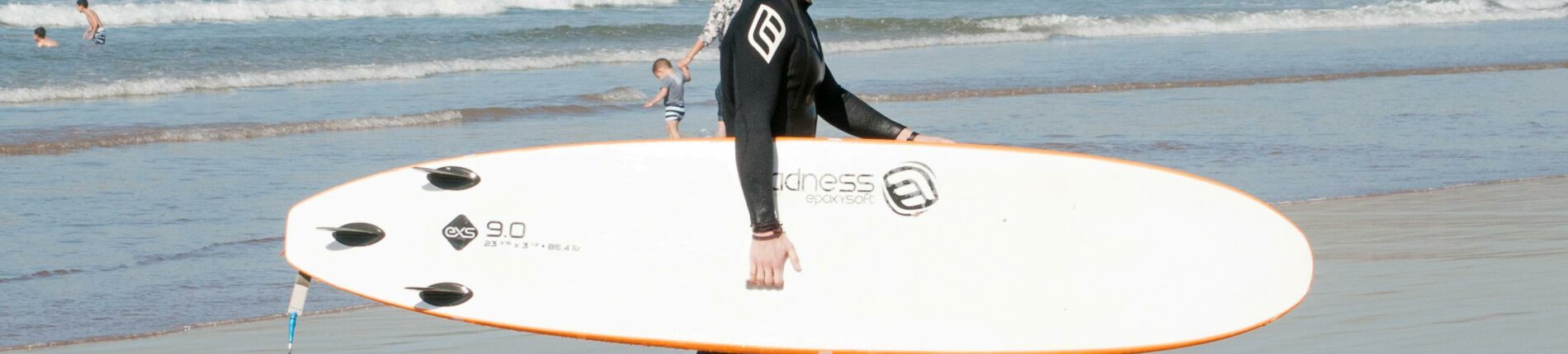 A surfer walks along Agadir beach carrying a surfboard, reflecting in wet sand.