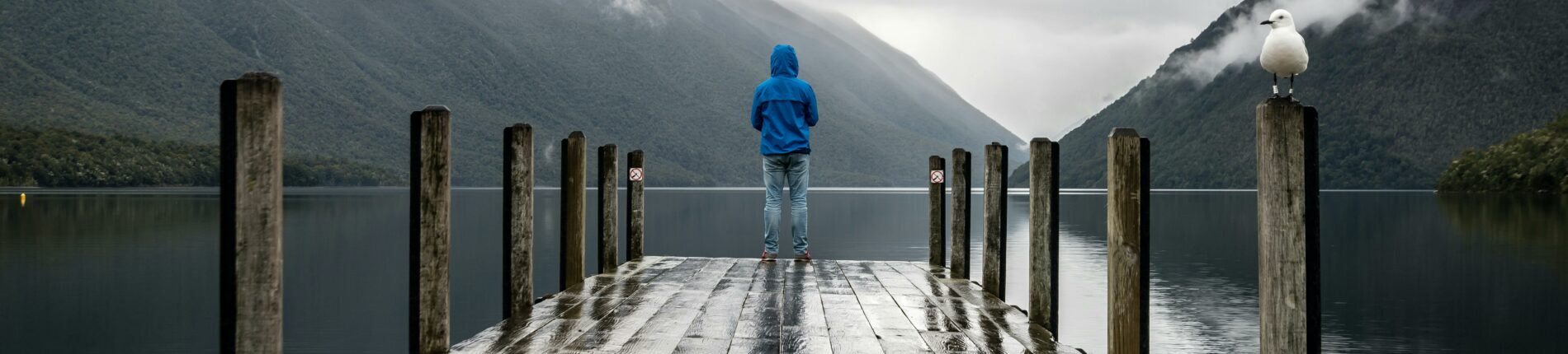 Person in blue jacket stands on a rainy dock overlooking serene lake and mountains.