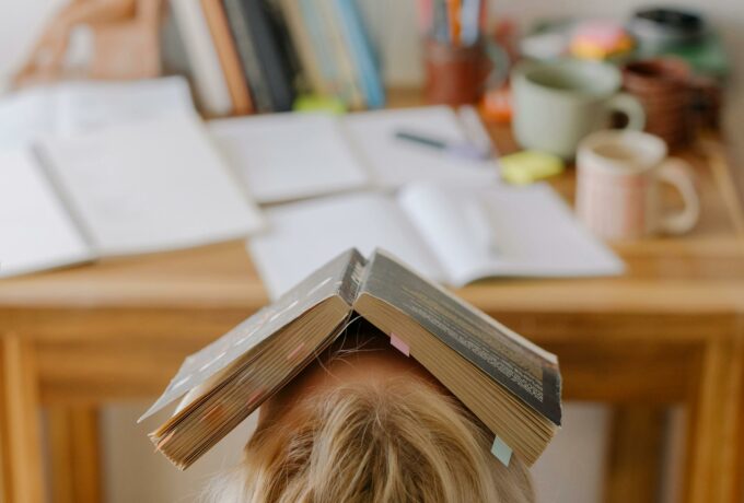 Student feeling stress and exhaustion while studying at a cluttered desk with an open book on their head.