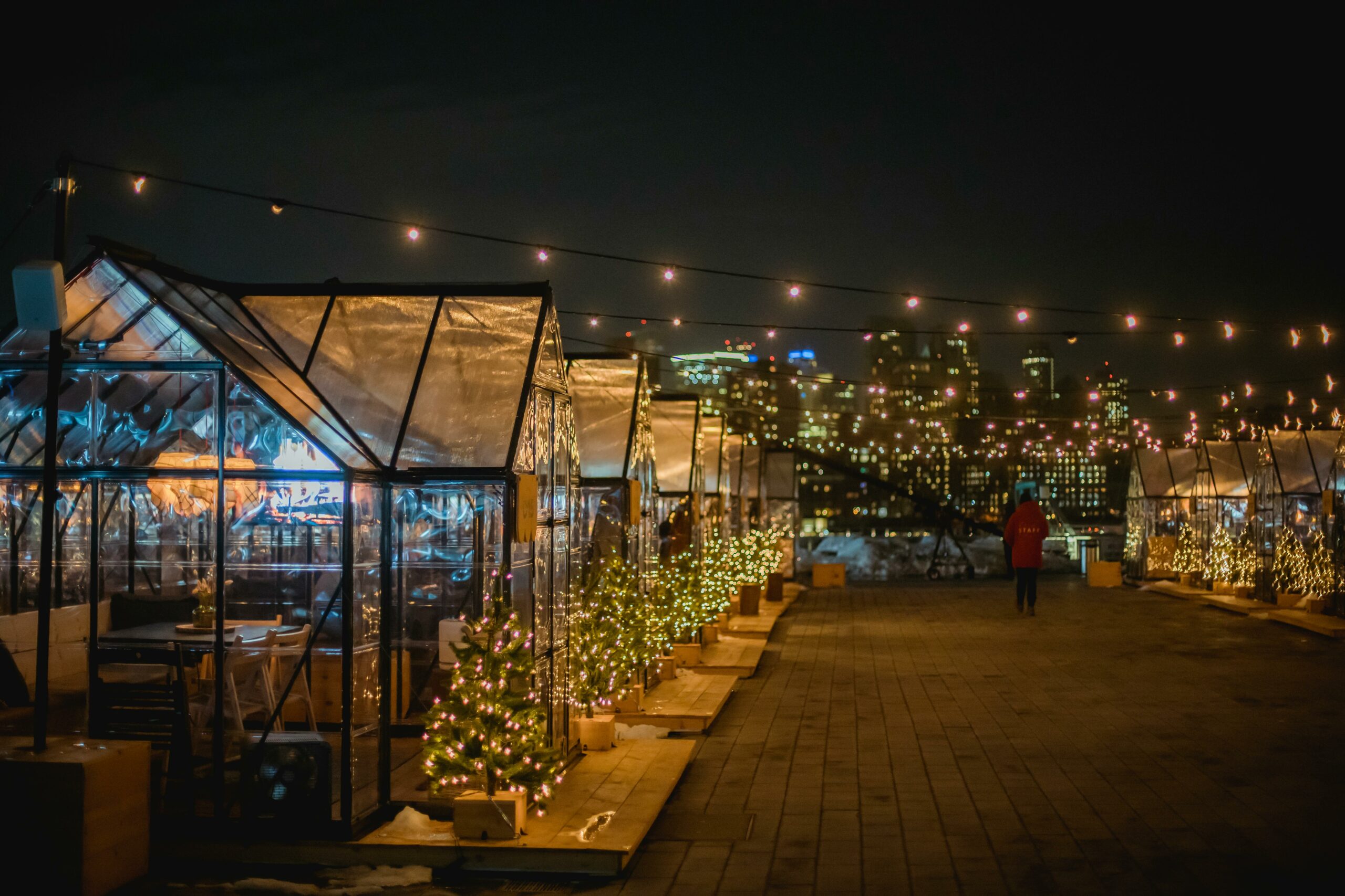 Glowing Christmas trees and festive lights adorn a rooftop market with cityscape views at night.