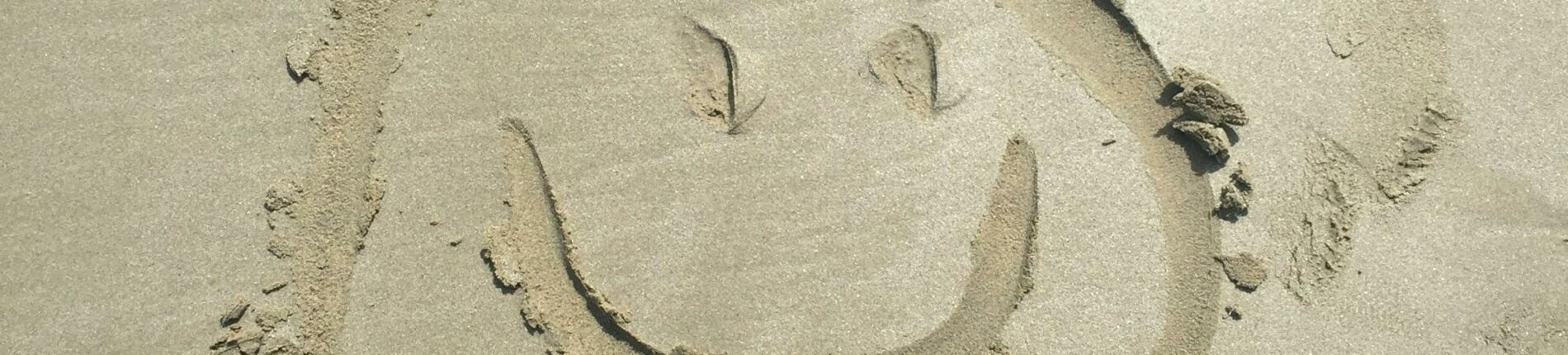 A smiley face drawn in sand on a sunny beach, captured with shoes visible.