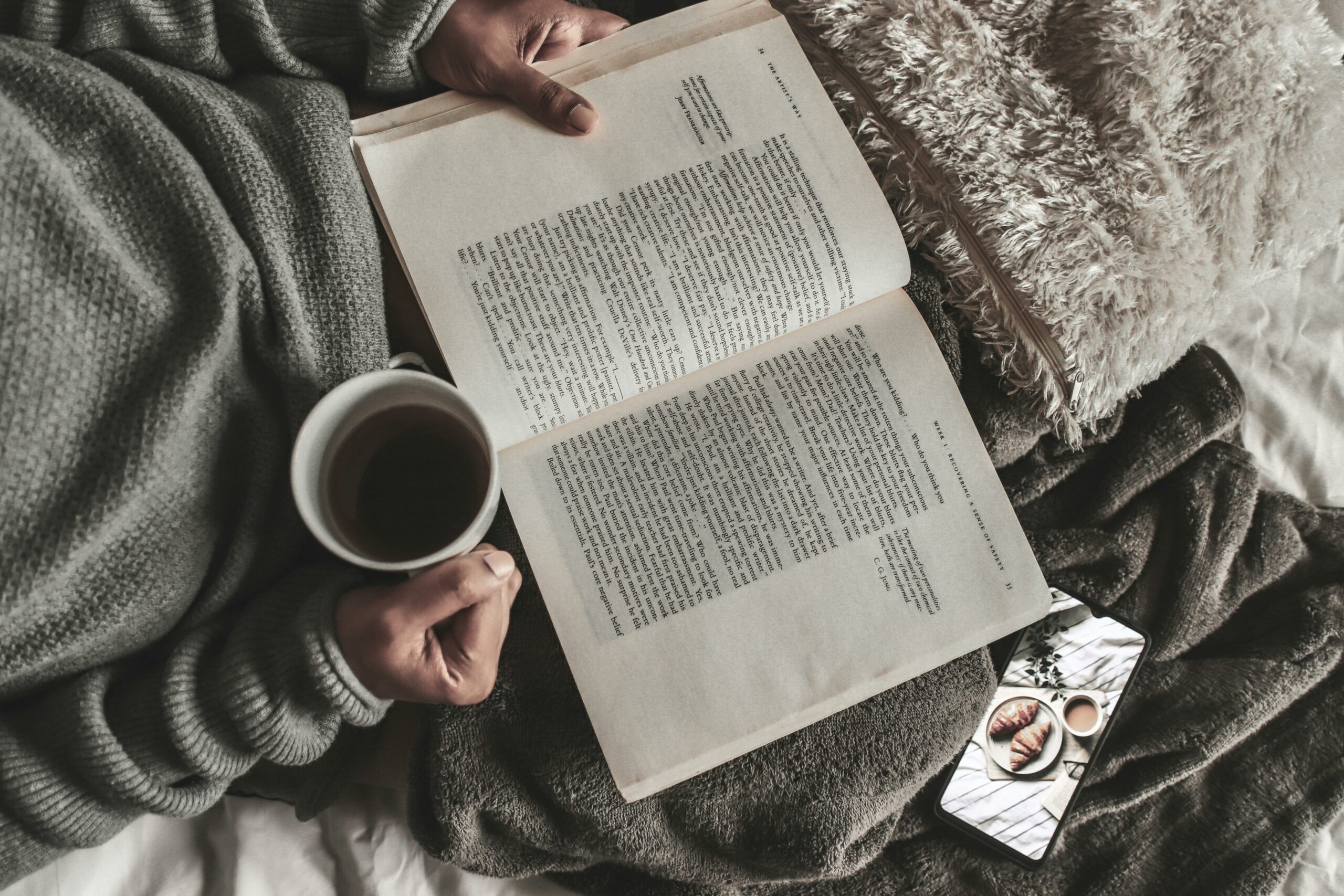 From above of crop anonymous female with mug of hot tea reading book under plaid near smartphone at home